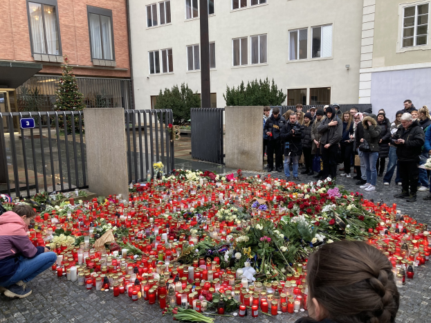 Eine Gruppe von Menschen an einer improvisierten Gedenkstätte mit Blumen und Kerzen vor einem Gebäude, eine Person kniet und hält eine Kamera; Gebäude, Pflanzen, ein Pfahl und ein Tor sind im Hintergrund zu sehen.