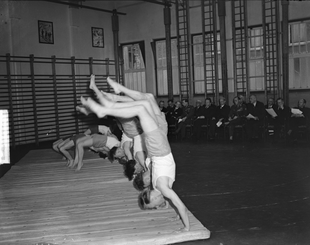 Eine Gruppe führt Akrobatik auf einer Bühne vor einem Publikum auf, mit Foto-Rahmen, Fenstern und Leitern im Hintergrund, dargestellt in Schwarz-Weiß.