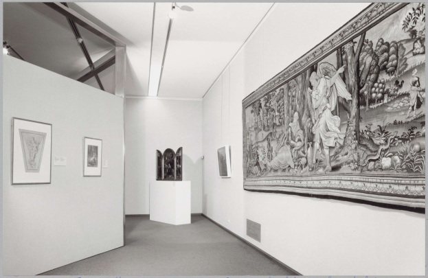 Schwarzes und weißes Foto eines Museumsraums mit einem großen Wandteppich, gerahmten Kunstwerken an den Wänden, Objekten auf einem Podest und Deckenleuchten, mit dem Text "Museum of Fine Arts, New York City" unten.