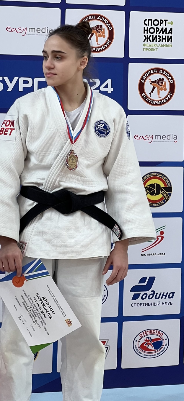 Frau in weißem Judo-Anzug mit schwarzem Gürtel auf einem Podest, die eine Medaille und ein Zertifikat hält, vor einem Hintergrund mit Logos und Text.