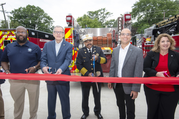 Eine Gruppe von Menschen schneidet eine rote Schleife vor einem Feuerwehrauto, feierlich zur Einweihung der Alexandria Fire Department, mit Bäumen, Polen, Drähten und einem klaren blauen Himmel im Hintergrund.