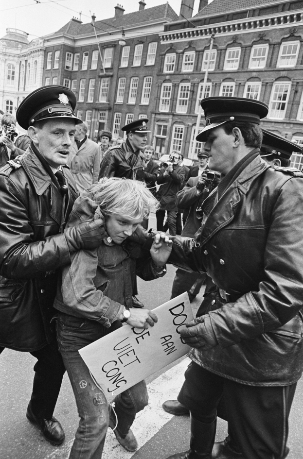 Schwarz-weiß-Foto einer Frau, die von zwei Polizeibeamten auf einer Straße eskortiert wird, mit Zuschauern und Gebäuden im Hintergrund.