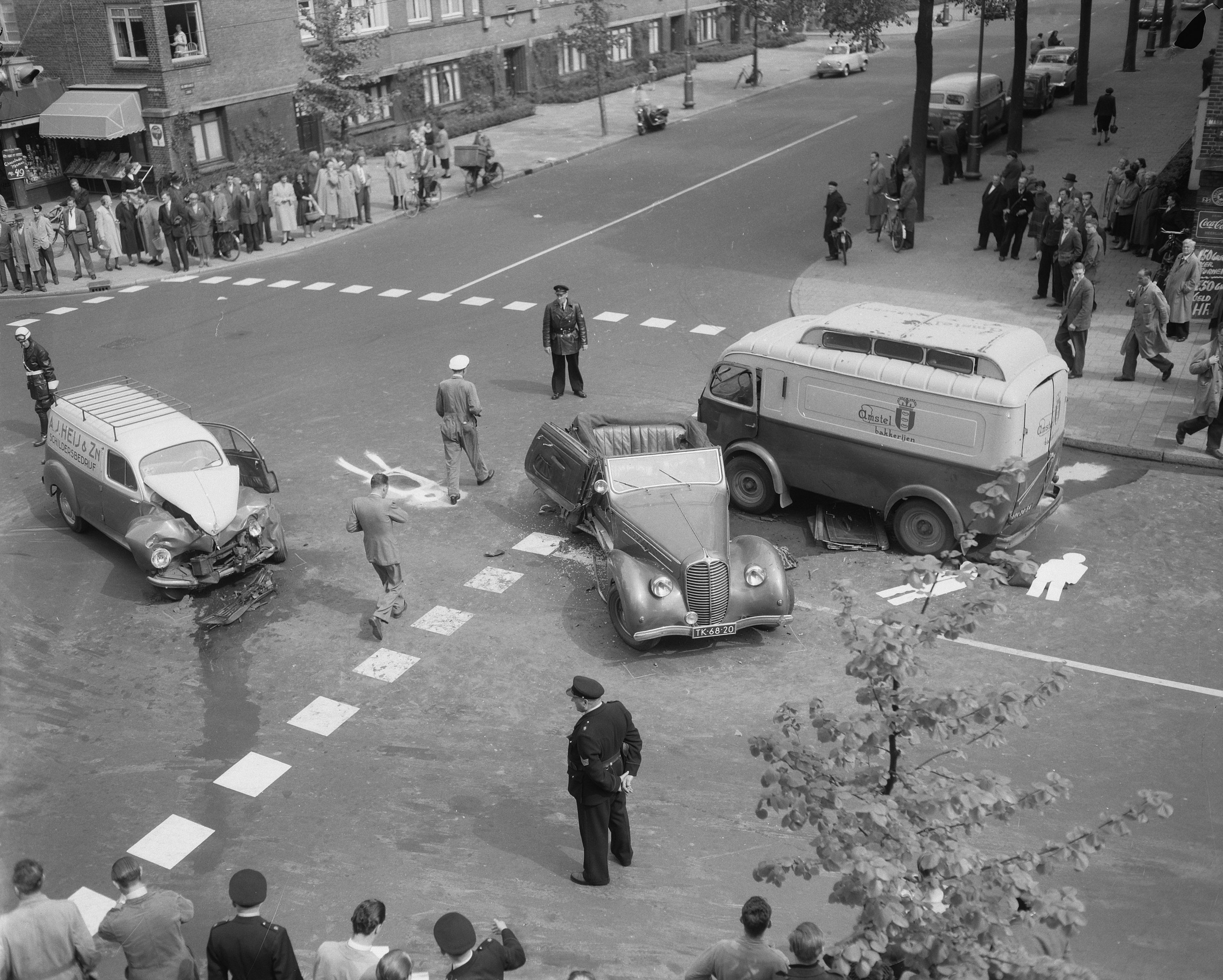 Schwarz-weißes Bild eines Autounfalls an einer Straßenkreuzung mit einem Van, anderen Fahrzeugen und Menschen, die sich darum versammelt haben, mit Gebäuden, Bäumen, Pfosten und Texttafeln im Hintergrund.