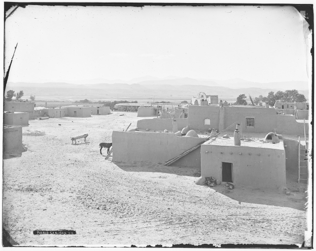 Schwarzes und weißes Foto einer Adobe-Dorfschaft in einer Wüstenlandschaft mit Häusern, Bäumen, Bergen im Hintergrund und Tieren am Boden, unter einem sichtbaren Himmel.