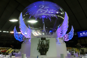 Ein großer Kristallball mit Flügeln oben drauf, umgeben von Blumengestecken, Namenschildern und anderen Gegenständen auf dem Boden, vor einem dekorierten Hintergrund mit Brettern, Lichtern und einem Bildschirm für ein 25-jähriges Jubiläum.