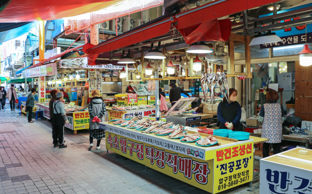 Ein belebter Straßenmarkt in Seoul, Südkorea mit Menschen, Ständen, die verschiedene Gegenstände verkaufen, und Gebäuden unter einem klaren blauen Himmel.