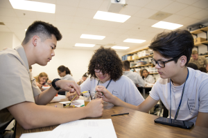 Three students sit at a table in a classroom, collaborating on a project with papers, pens, and a mobile phone visible on the table, while racks, lights, and a projector are seen in the background.