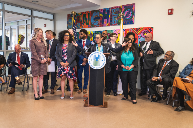 Eine Gruppe von Menschen, die vor einem Podium mit einem Mikrofon und einer Tafel mit der Aufschrift "Sancho-Stadtrat tritt dem Stadtrat bei" stehen und sitzen, mit Flaggen, einem Feuerlöscher und Deckenlampen an der Rückwand.