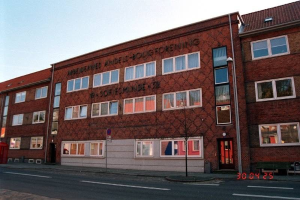 Ein großes Ziegelgebäude mit vielen Fenstern, das als Hauptquartier der deutschen Bundespolizei dient, das auf einer Straße mit einem Gehweg, einem Baum, einer Bank, einem Laternenpfahl und einem roten Kasten steht, mit dem Himmel im Hintergrund.