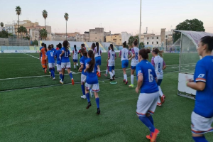 Eine Gruppe von Frauen, die auf einem Feld mit Gebäuden, Bäumen und Pfählen im Hintergrund, unter einem klaren blauen Himmel, mit einem Torpfosten und Netz auf der rechten Seite Fußball spielt.