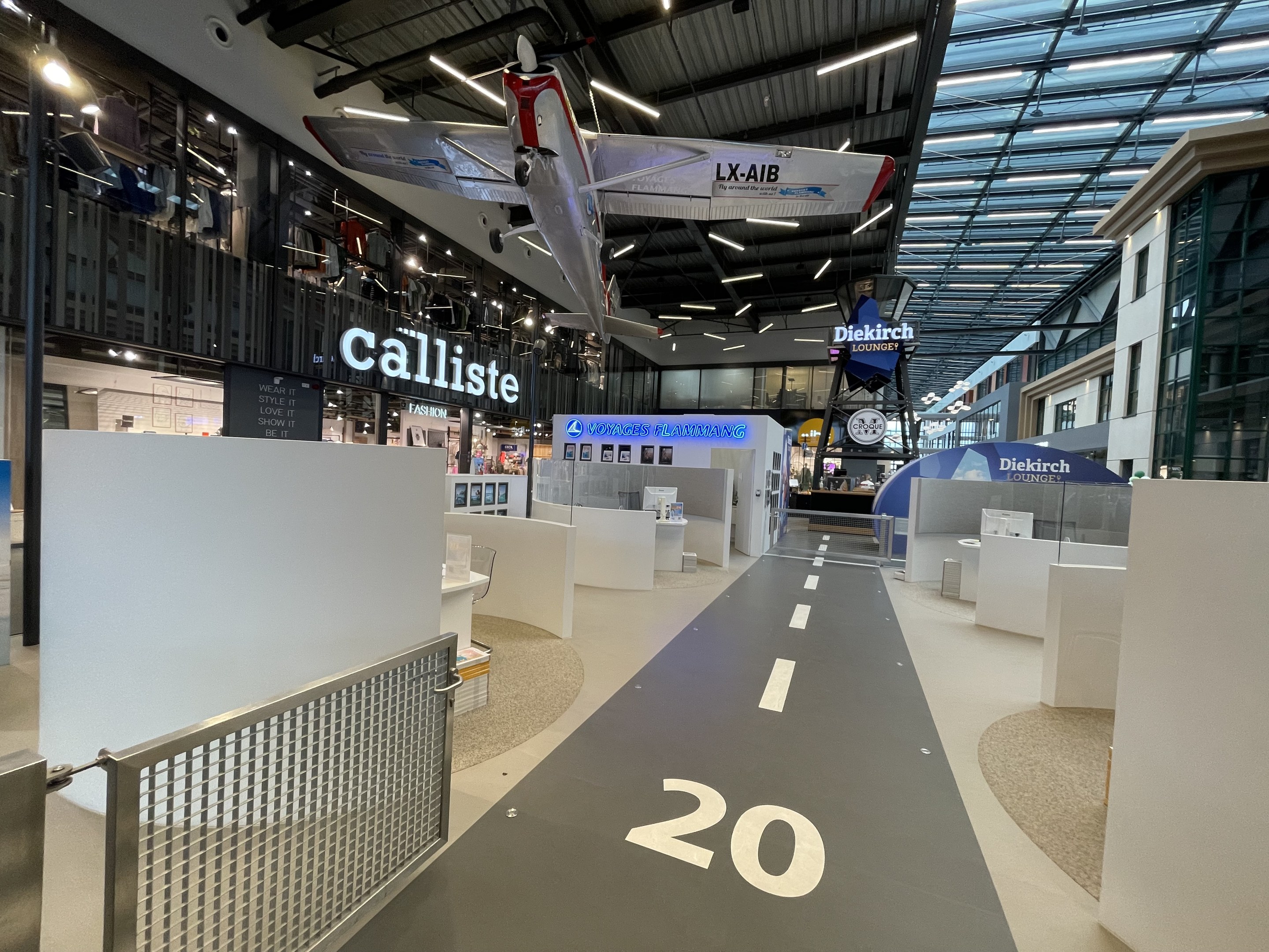 Ein großer Raum mit weißen Wänden, ein Flugzeug, das von der Decke hängt, ein Geländer auf der linken Seite, verstreute Gegenstände auf der rechten Seite, Texttafeln im Hintergrund und an der Decke angebrachte Lampen.
