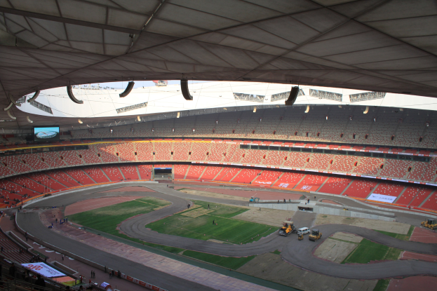 Großes Stadion voller Menschen, Fahrzeuge auf der Straße, grasbewachsener Boden, Texttafeln, Geländer, ein Bildschirm und Deckenbeleuchtung.