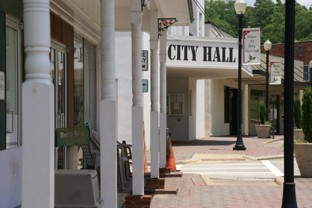 Rathausgebäude mit umliegender Infrastruktur, darunter Straßenmasten, Lichter, Namensschilder, Fahrzeuge, Verkehrskegel, Stühle, Topfpflanzen, Bäume und eine bewölkte Himmel.
