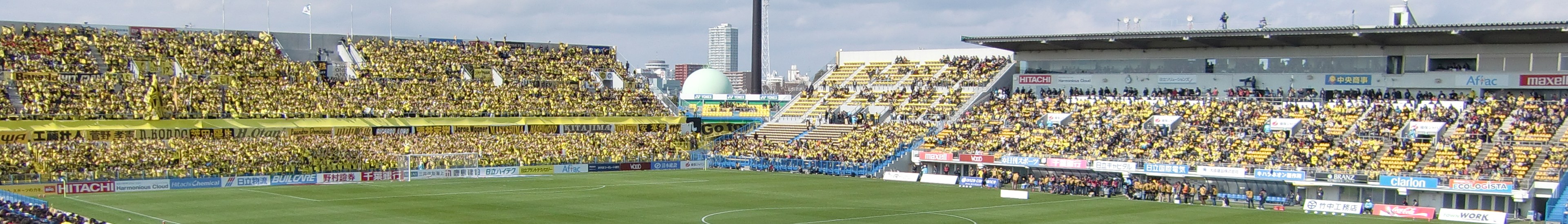 Großes Stadion voller Zuschauer bei einem Fußballspiel auf saftigem Grün mit Umrandungstafeln, Gebäuden, Pfählen und einem klaren blauen Himmel im Hintergrund.