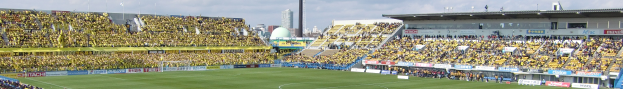 Großes Stadion voller Zuschauer bei einem Fußballspiel auf saftigem Grün mit Umrandungstafeln, Gebäuden, Pfählen und einem klaren blauen Himmel im Hintergrund.