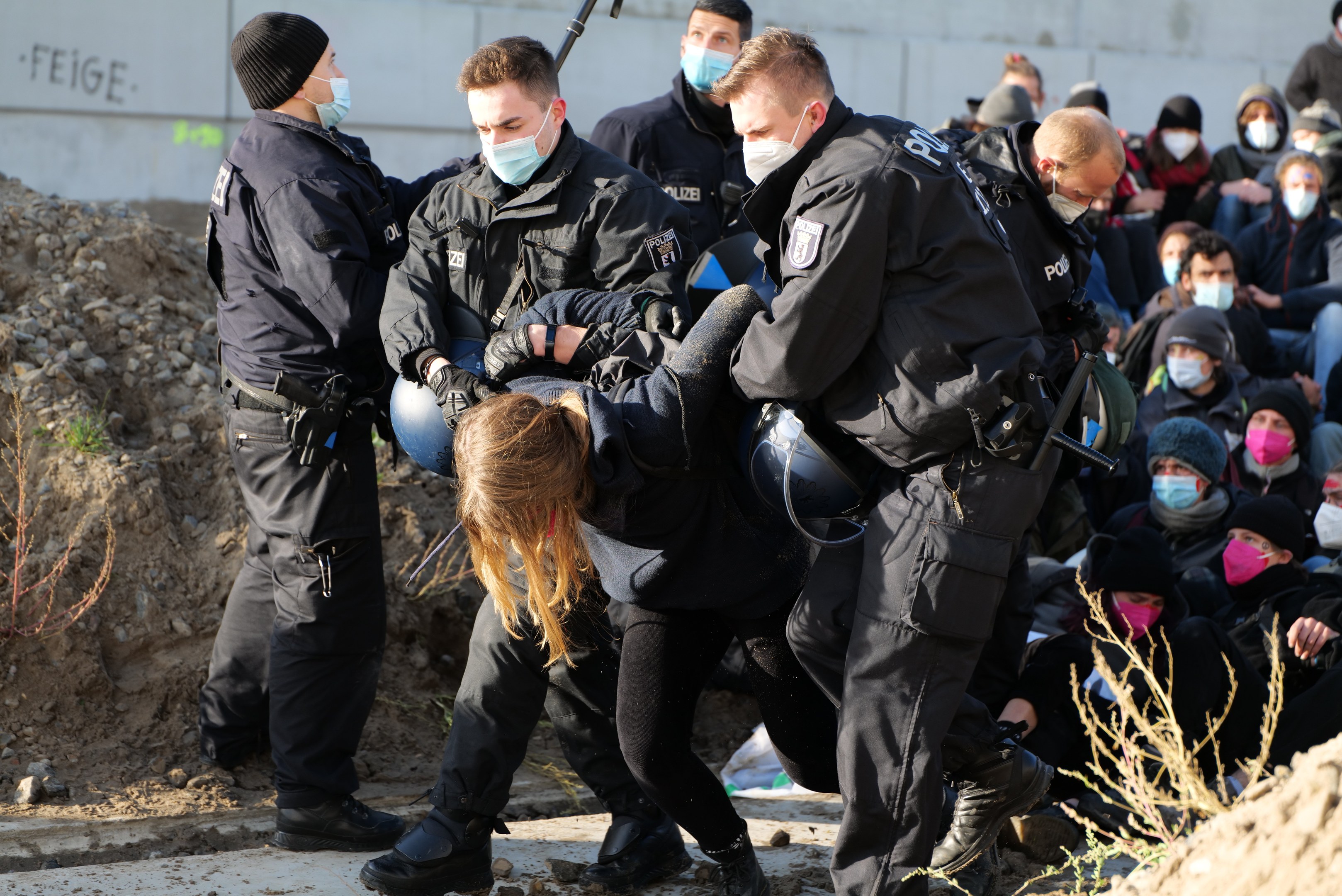 Gruppe maskierter Menschen in Mützen, einige mit Waffen, umgeben eine Frau, die von der Polizei eskortiert wird, in einer städtischen Umgebung mit einer Wand mit Text und Pflanzen.