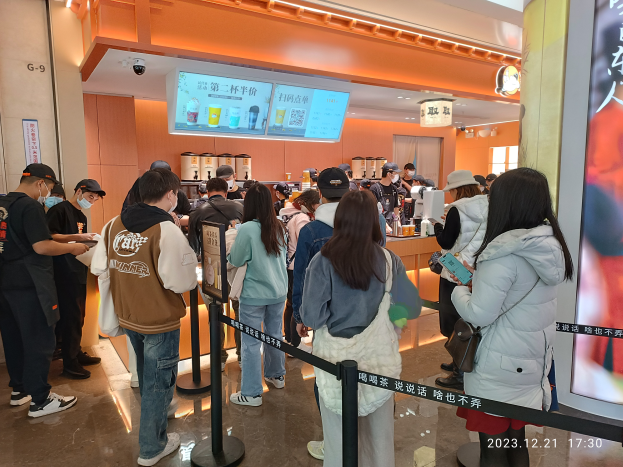 Eine Gruppe von Menschen steht an einem Tresen in einem Restaurant Schlange, einige tragen Mützen und Taschen, andere halten Gegenstände in den Händen, mit Stangen, Bändern, einem Bildschirm, Deckenleuchten und Tischen mit verschiedenen Gegenständen im Hintergrund.