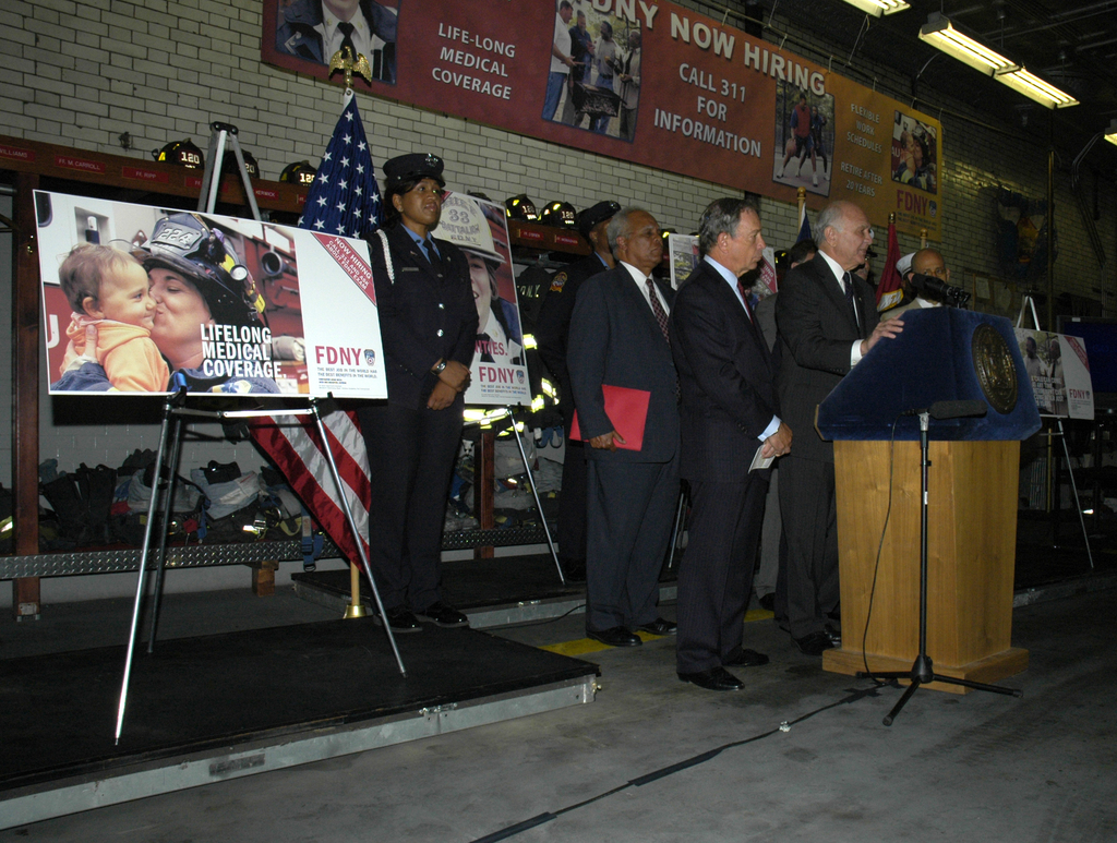 Eine Gruppe von Menschen steht in der Nähe eines Podiums mit Mikrofonen, Schildern auf Ständern, Fahnen und Schildern an der Wand, alles unter hellen Scheinwerfern.