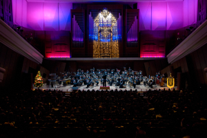 Ein großer Saal voller Menschen, die ein Konzert genießen, mit Musikern auf der Bühne, einem Weihnachtsbaum im Hintergrund und Wänden, die mit Lichtern und anderen Gegenständen geschmückt sind.