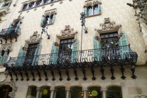 Ein Gebäude mit Fenstern, Balkonen mit Pfosten und Wandreliefs, mit einem Kronleuchter im Inneren am unteren Bildrand sichtbar.