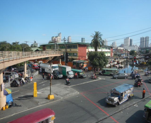 Eine belebte Stadtstraße mit Autos, Bussen, Motorrädern, einer Brücke mit Geländern und Säulen, Laternen, Strommasten mit Drähten, Gebäuden mit Fenstern, Bäumen und einem bewölkten Himmel.