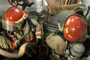 Feuerwehrleute in Schutzausrüstung an einem Feuerhydranten mit Rohren und Ventilen im Hintergrund.