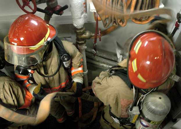 Feuerwehrleute in Schutzausrüstung an einem Feuerhydranten mit Rohren und Ventilen im Hintergrund.