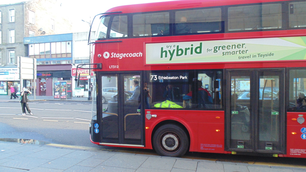 Ein Zahlungsterminal und ein Bus im Vordergrund mit Gebäuden, Gehweg, Werbetafeln, Menschen und einer Straße auf der linken Seite unter einem sonnigen Himmel.