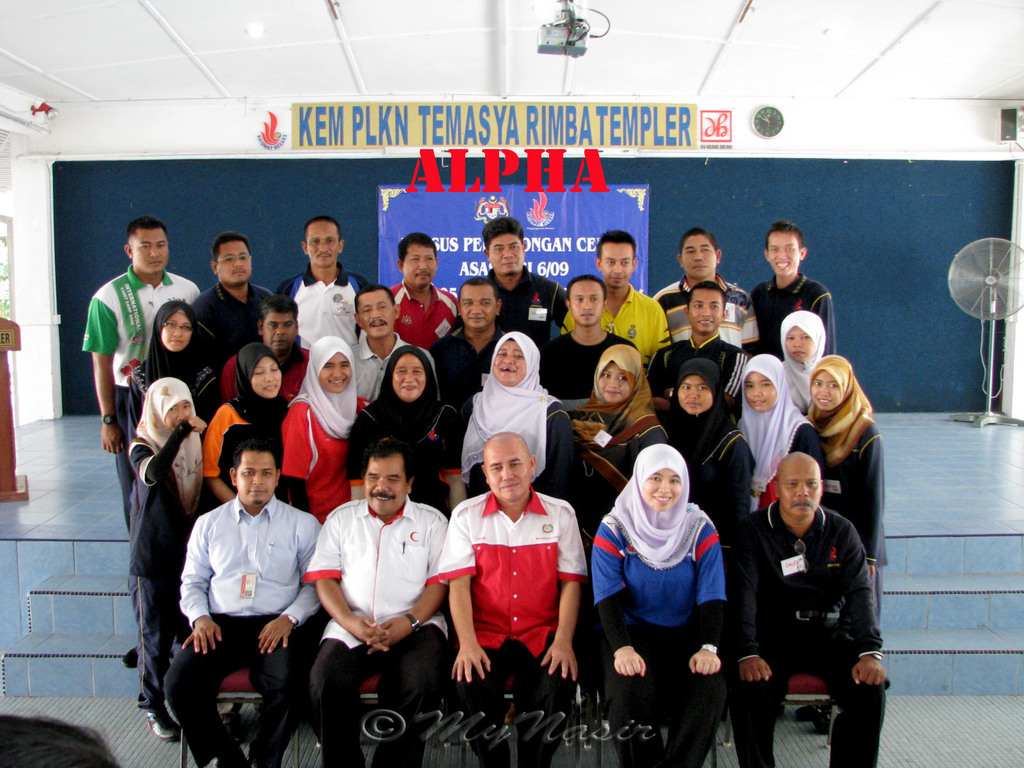 Eine Menschenmenge, die lachend vor einer Bühne posiert, mit einer Tafel mit Text an der Wand dahinter, einem Tischventilator rechts, einem Podium links und einem Projektor oben im Bild.