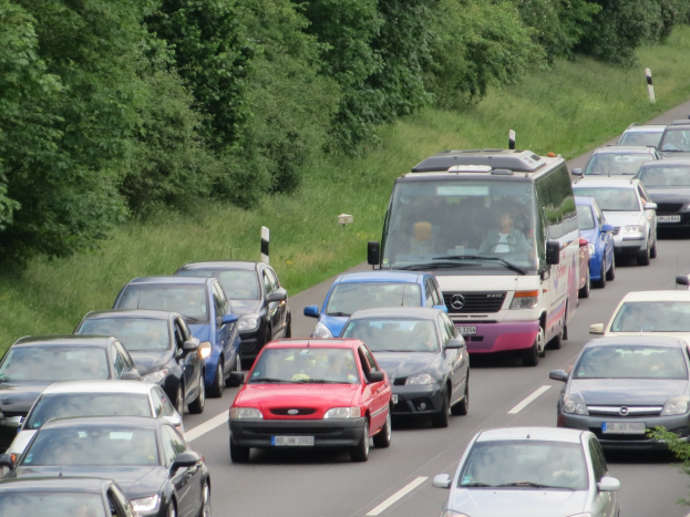 Ein Stau auf einer Autobahn mit mehreren Autos und einem Lieferwagen, Menschen sichtbar in den Fahrzeugen, Bäume und Gras im Hintergrund.