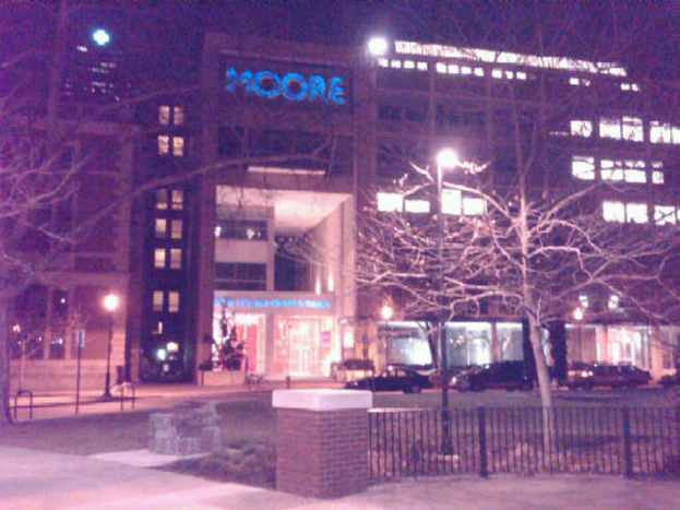 Außenansicht eines gebäudeähnlichen Krankenhauses mit parkenden Fahrzeugen, Bäumen und einem Zaun im Vordergrund.