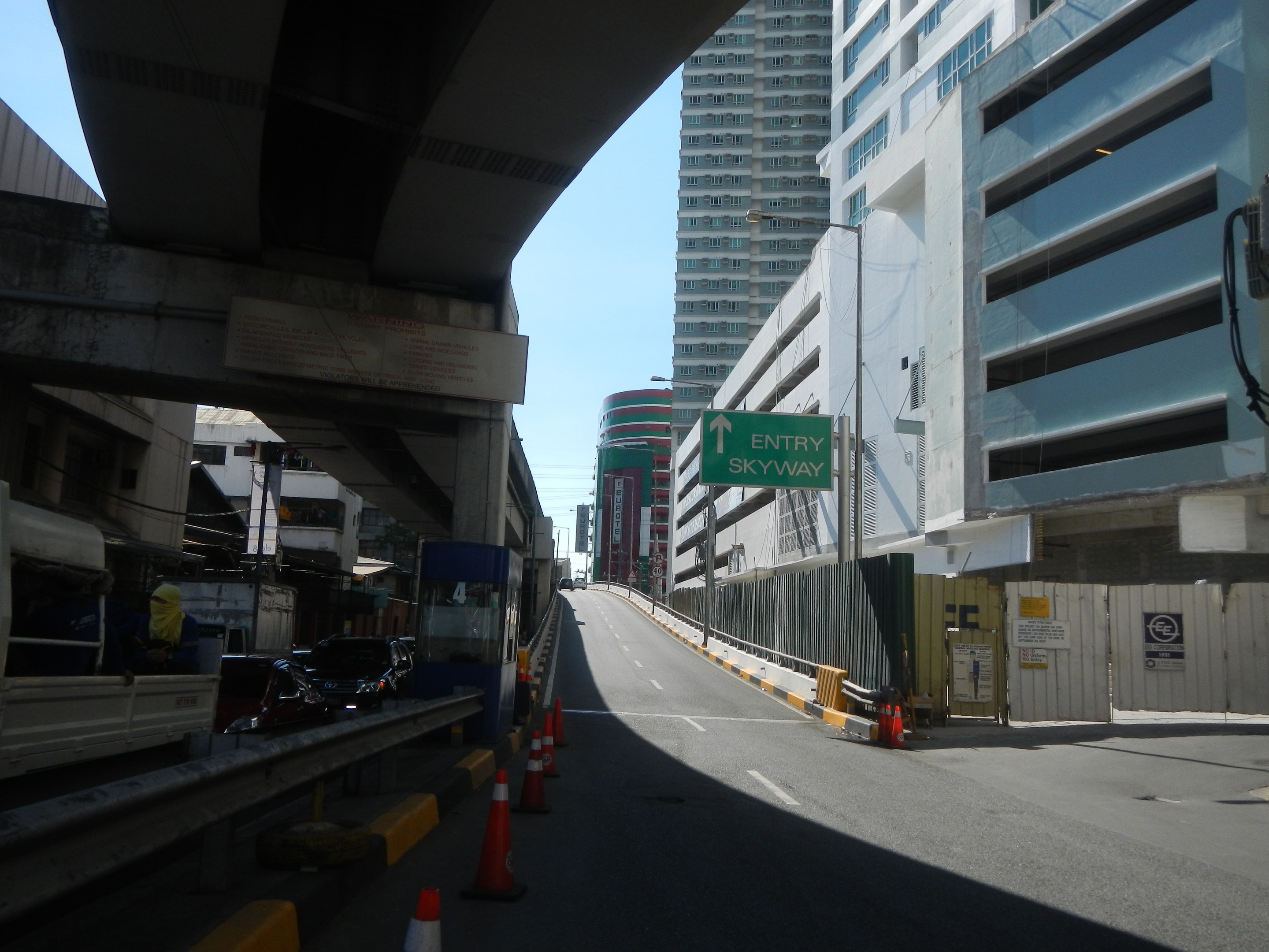Stadtstraße mit Verkehrskegeln, Fahrzeugen, einer Brücke auf der linken Seite, Gebäuden mit Namensschildern, Laternen und einem Himmel im Hintergrund.