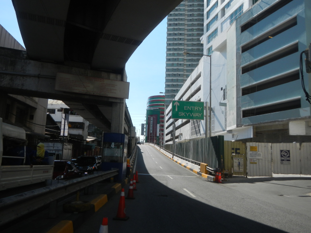 Stadtstraße mit Verkehrskegeln, Fahrzeugen, einer Brücke auf der linken Seite, Gebäuden mit Namensschildern, Laternen und einem Himmel im Hintergrund.