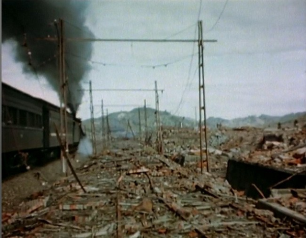 Ein Zug fährt durch ein beschädigt Gebiet mit Rauch, der aus ihm aufsteigt, umgeben von Strommästen und -drähten, Hügel im Hintergrund bei einem klaren Himmel, mit verstreuten Trömmern am Boden.