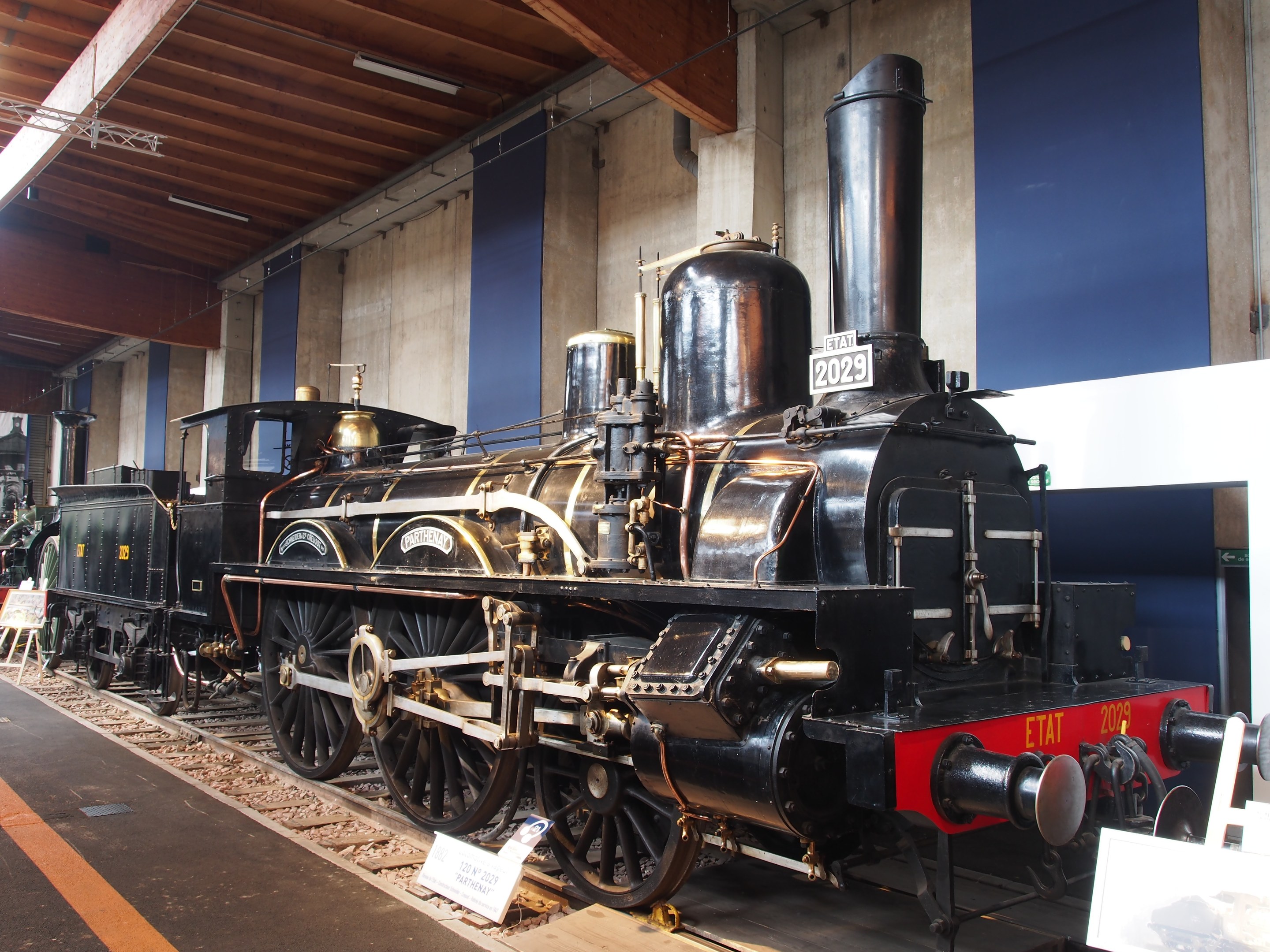 Eine große, alte Dampflokomotive, die in einem Museums Setting ausgestellt ist, umgeben von Eisenbahnschienen, Informationsschildern und anderen Ausstellungen, mit einer beleuchteten Decke und Wand im Hintergrund.