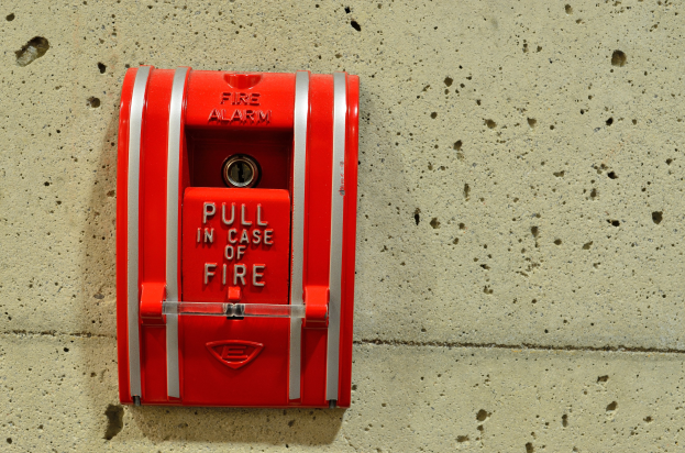 Roter Feueralarm an einem Gebäude montiert, zylindrischer Metallkasten mit Griff und Schlüsselloch, beschriftet mit "Feueralarm" in schwarzer Schrift.