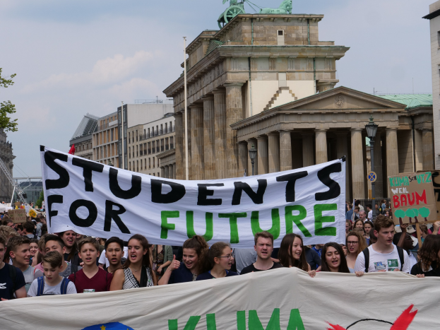 Gruppe von Schülern marschiert in Berlin mit einer bunt bemalten "Students for Future"-Schleife vor einer Kulisse aus Gebäuden, Bäumen und Himmel.