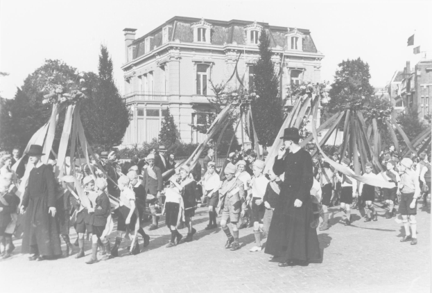 Schwarz-weißes Bild einer Gruppe von Menschen, die die Straße entlanggehen und Fahnen halten, mit Hüten, Bäumen, Gebäuden und einem Maibaum im Hintergrund.