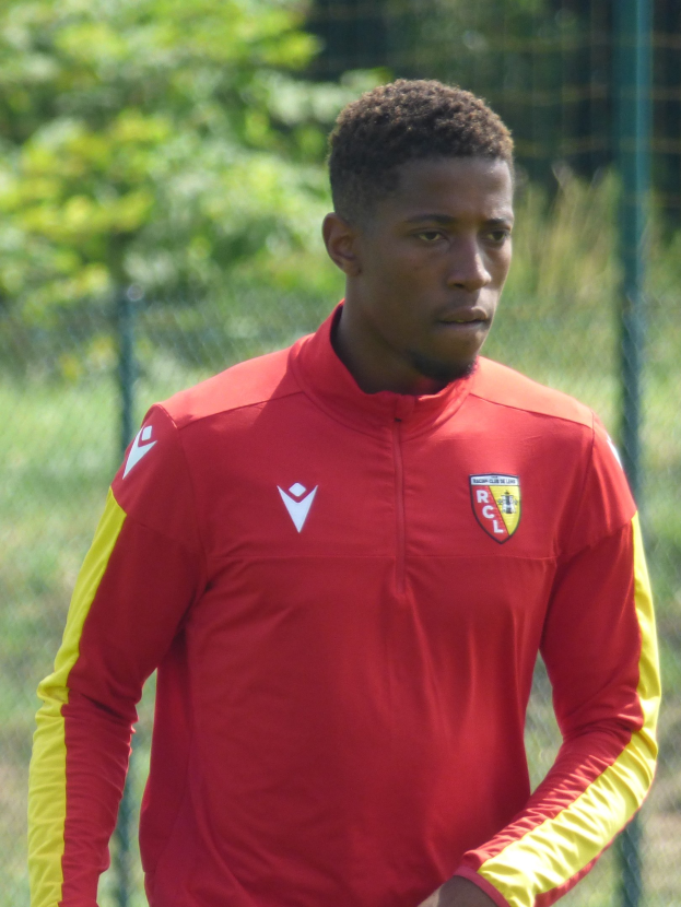 Ein junger Mann in rot-gelbem Trikot steht auf einem Fußballfeld umgeben von einem Zaun und Bäumen und schaut entschlossen.