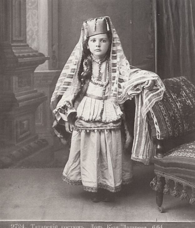 Schwarzes und weißes Foto eines jungen Mädchens in traditioneller russischer Kleidung, das rechts neben einem Stuhl steht, mit einem Vorhang und einer Wand im Hintergrund und Text unten.