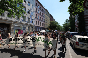 Eine Gruppe von Menschen marschiert auf einer Straße und hält eine Fahne hoch, mit parkenden Autos auf der rechten Seite, einem Radfahrer auf der linken Seite und Gebäuden, Bäumen und einem Schild im Hintergrund unter einem sichtbaren Himmel.