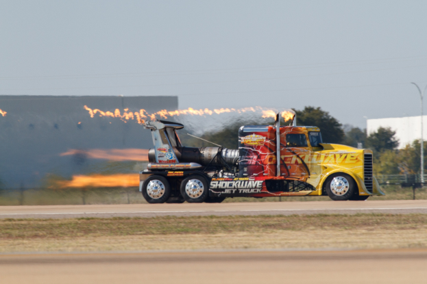 Flammen schlagen aus dem Heck eines Lkw, der auf einer von Gras, einem Zaun, B├Ąumen, einem Geb├Ąude, einem Laternenpfahl, Dr├Ąhten und einem bew├Âlktem Himmel ges├Ąumten Stra├če fahrenden Trucks.