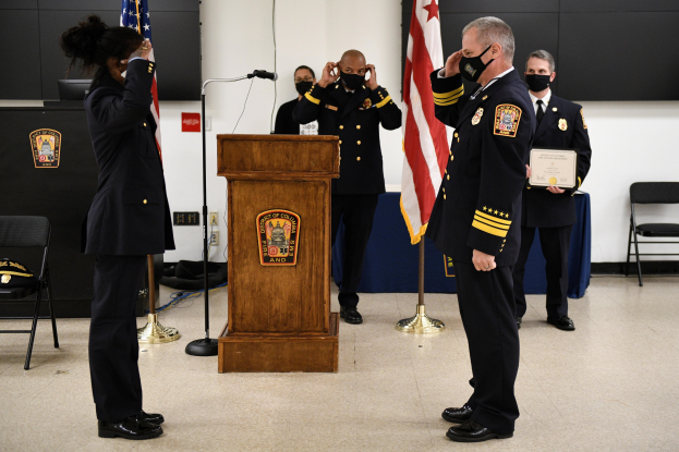 Gruppe uniformierter Personen in Masken auf einem Podium während einer Vereidigung eines neuen Feuerwehrchefs, der ein Zertifikat hält, mit Flaggen, Stühlen, Tischen und einer Wand im Hintergrund.