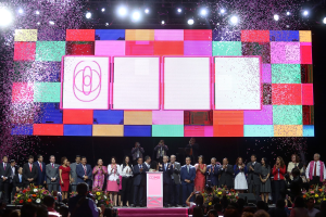 Gruppe von Menschen auf einer Bühne mit einem Podium, Blumengestecken und Kameras, mit einem großen Bildschirm und Lichtern im Hintergrund während der Eröffnungszeremonie des China International Film Festivals 2018.