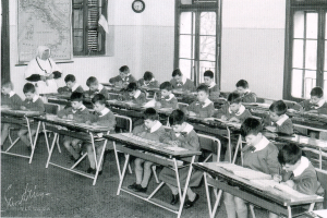 Ein Schwarz-Weiß-Foto einer Klasse mit Kindern an Tischen, Bücher und Papiere auf den Tischen, eine Uhr und eine Landkarte an der Wand und Fenster im Hintergrund.
