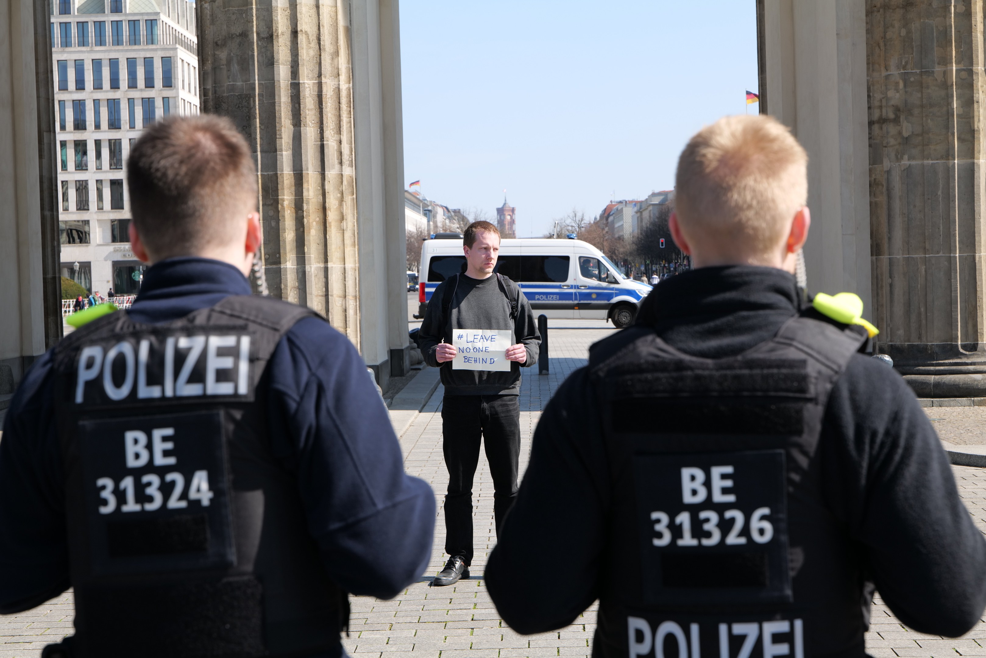 Drei Polizeibeamte stehen vor einem Gebäude mit einem Mann in der Mitte, der ein Papier hält, flankiert von Säulen, während Fahrzeuge, Menschen, Bäume und Gebäude den Hintergrund und den Himmel ausfüllen.