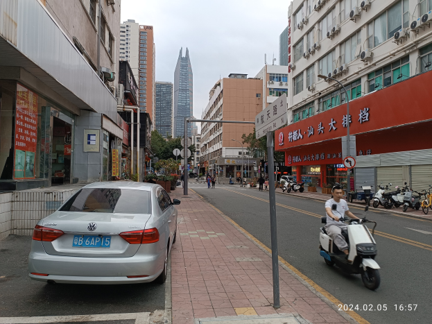 Ein Mann fährt auf einem Scooter eine von hohen Gebäuden gesäumte Stadtstraße entlang, mit Fußgängern auf den Gehwegen und anderen Fahrzeugen auf der Straße, unter einem klaren blauen Himmel mit Bäumen im Hintergrund.