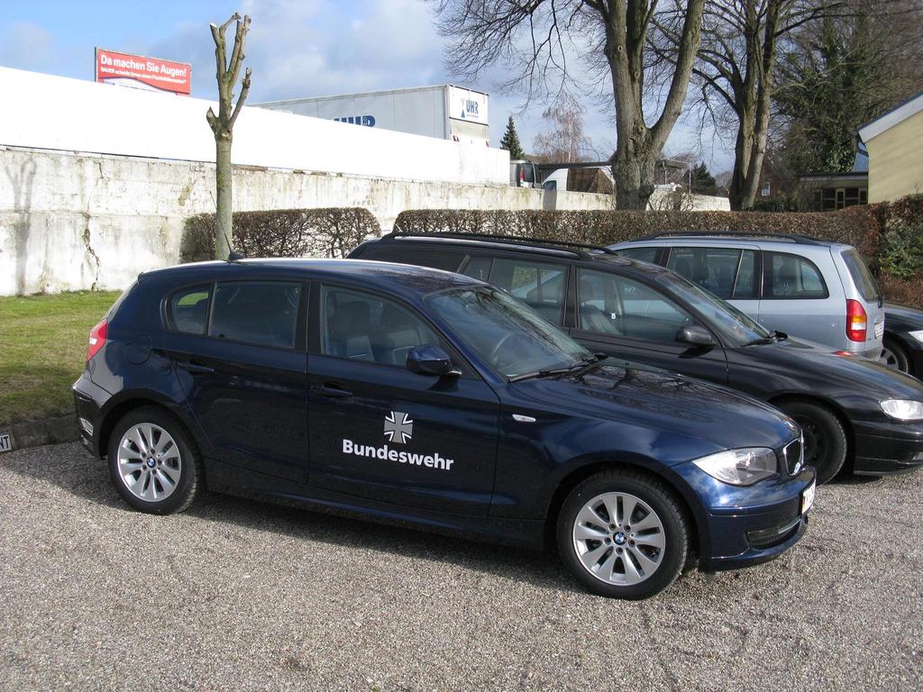Mehrere Autos auf einem Weg umgeben von grünem Gras, mit Büschen, Bäumen und Gebäuden im Hintergrund, ein Schild mit Text und ein Logo sichtbar.