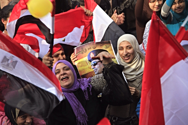 Demonstranten auf dem Tahrir-Platz in Kairo mit ägyptischen Flaggen und Parolen, einige mit Tüchern und Schildern, darunter eine Person mit einer Tafel und einem Luftballon im Hintergrund.