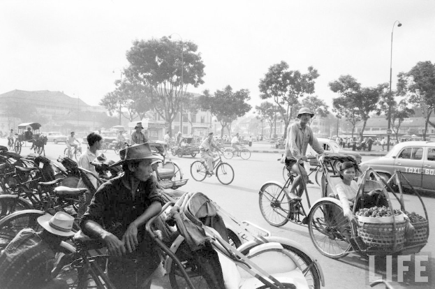 Eine viel befahrene Straße mit Fahrrädern, Autos und anderen Fahrzeugen, die an Bäumen und Gebäuden im Hintergrund vorbeifahren.